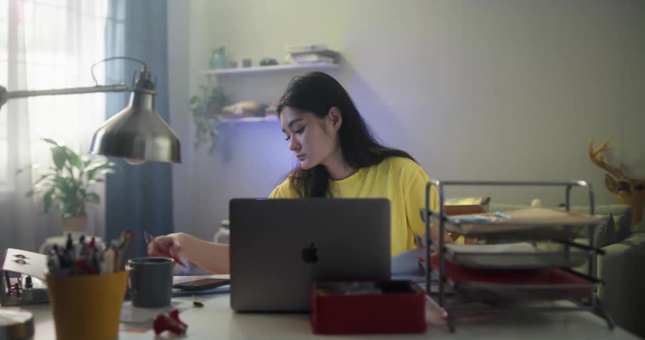 Asian girl looking very tired while studying on her notebook and laptop at the desk in the living room