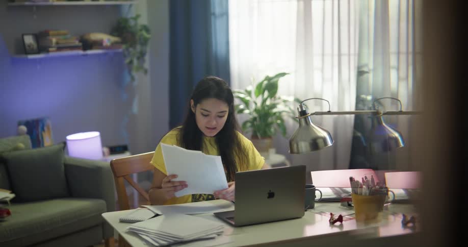 Teenage asian girl sitting at the desk having a video chat on her laptop while holding a stack of papers