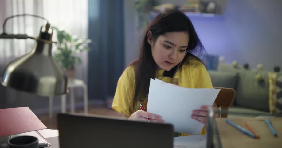 Teenage asian girl sitting at the desk talking to the phone while writing down and looking for something in a stack of papers
