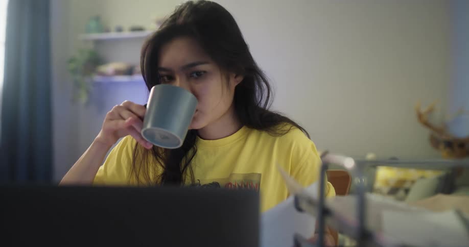 Tired asian girl sitting at the desk drinking a cup of coffee and studying on her laptop