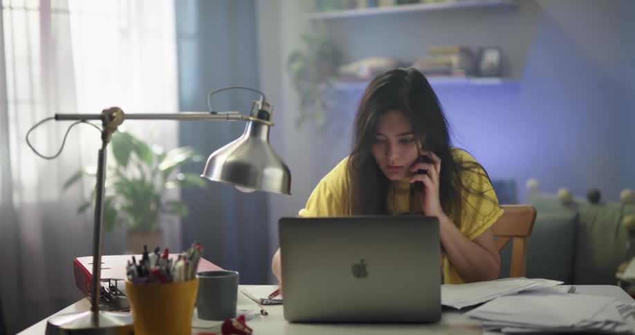 Young asian woman sitting at the desk talking on the phone while using her laptop and looking for something on some papers