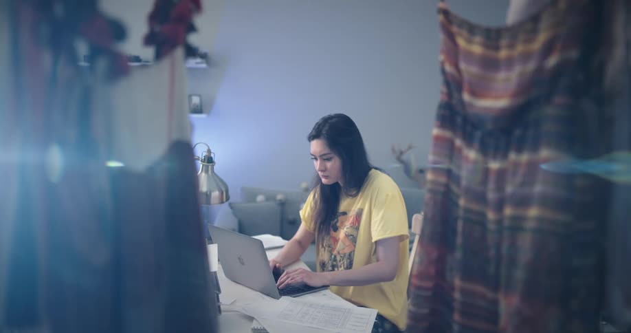 Woman with straight hair and blue and yellow pajamas works on lapton and answers call on smartphone