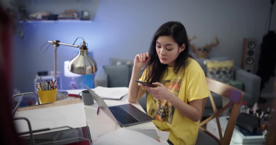 Busy and bored woman with dark straight hari and yellow t shirt sits at desk talking on smartphone and using desktop