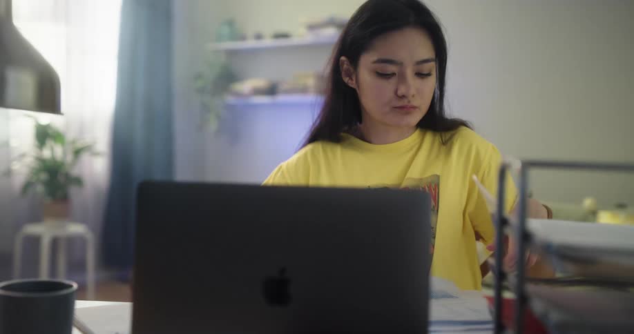Teenage asian girl sitting at the desk drinking a cup of coffee and studying with her laptop and some papers