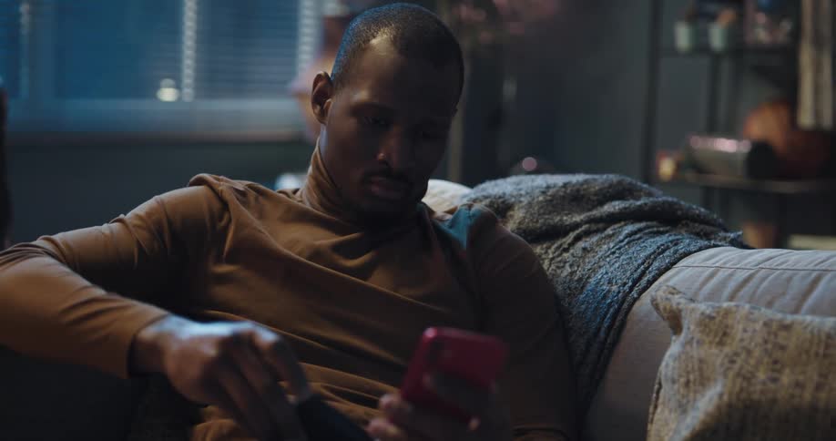 Man sitting on the couch with his legs up using his smartphone then taking a card from his pocket