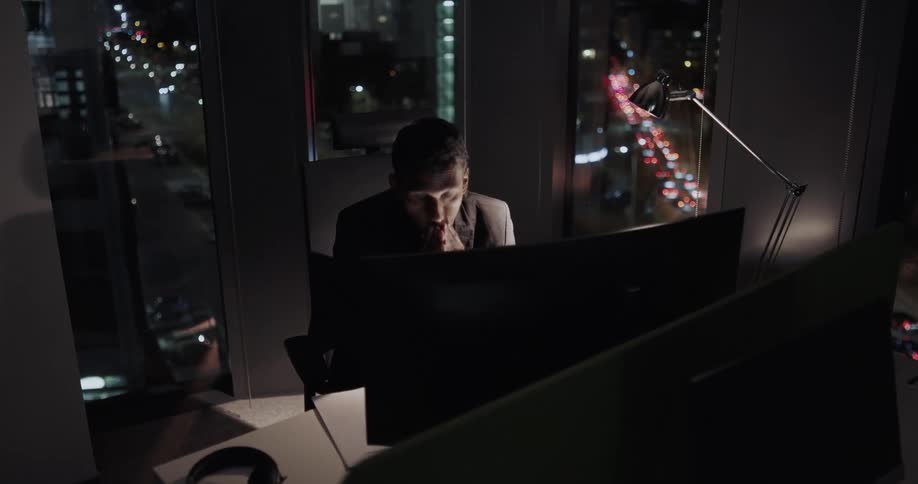 Male office worker sitting on his office space at night next to a big glass window looking very tired