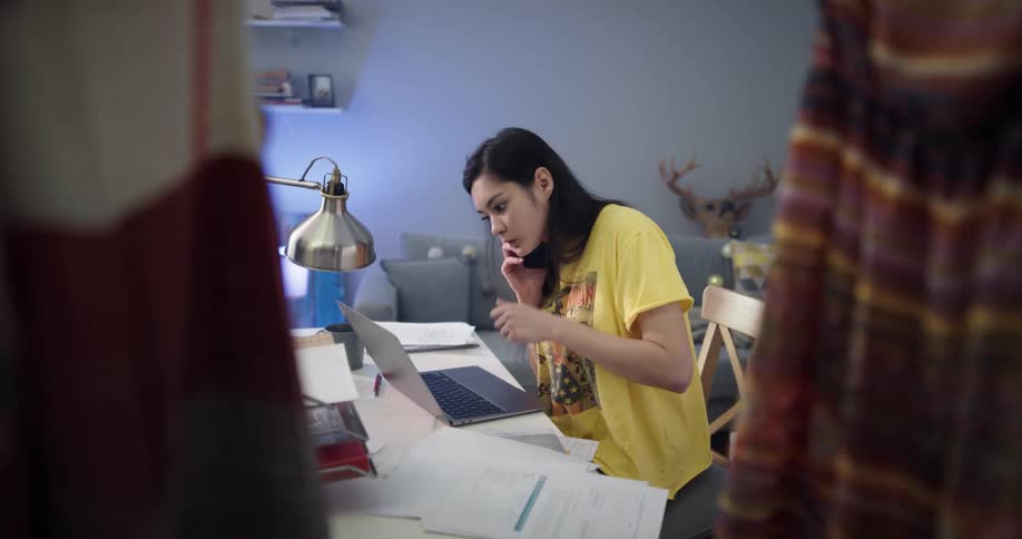 A young woman typing on a laptop stops to search for her phone before answering it and continuing to type