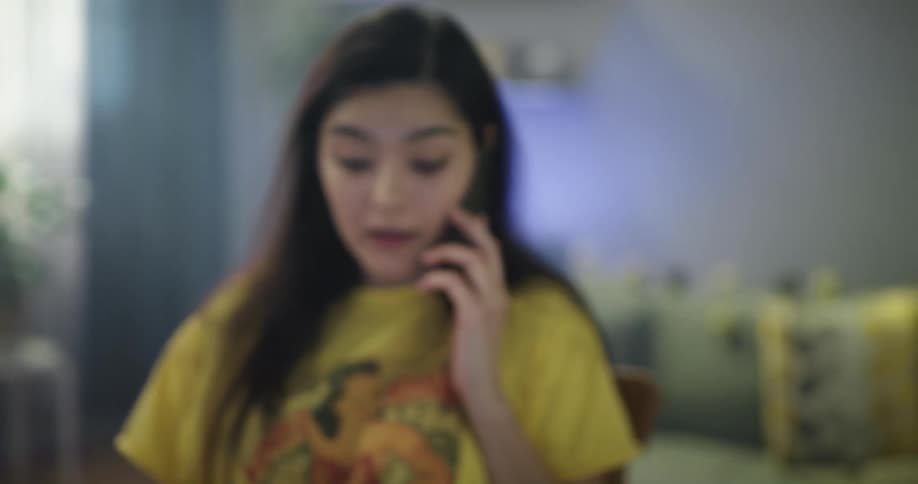 Young woman wearing yellow shirt working from home talks on the phone while checking paperwork