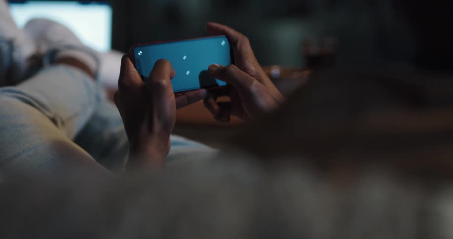 Man with jeans and shorts lies on couch and plays game on smartphone