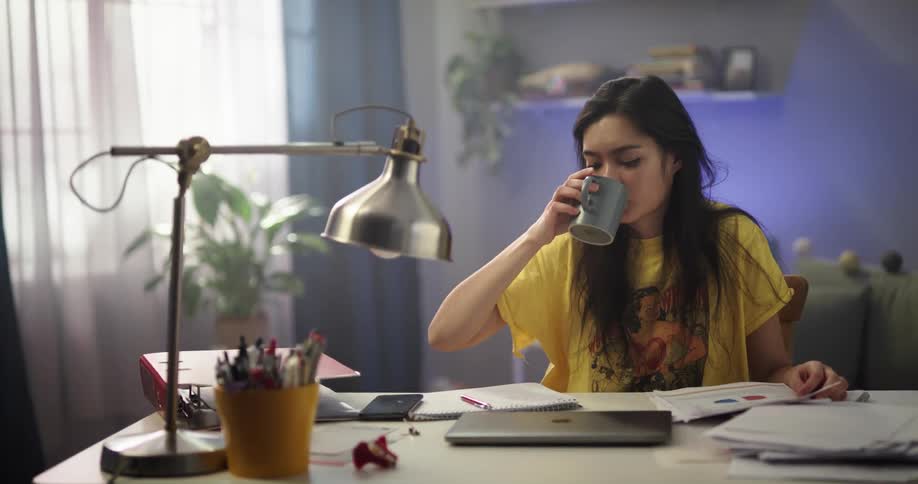 Tired and bored woman with long straight hair yellow and blue pajamas sits at desk closing laptop and drinking from cup