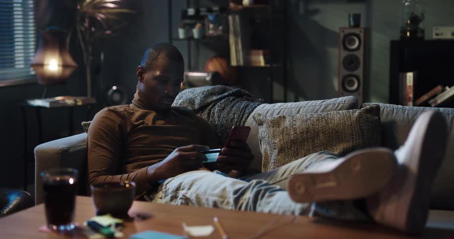 Man sitting on the couch with his legs up on the center table using his smartphone and grabbing his credit card from his pocket