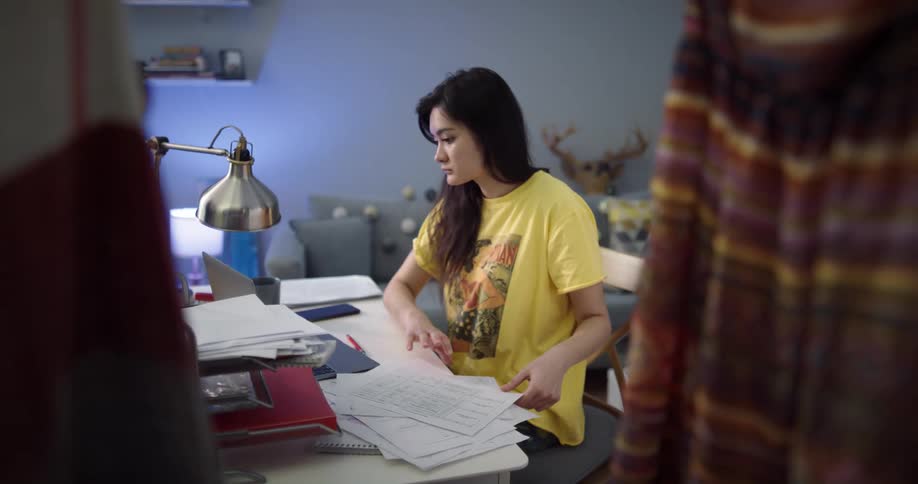 Young woman sitting at a desk in her room takes notes and organizes paperwork in front of a laptop