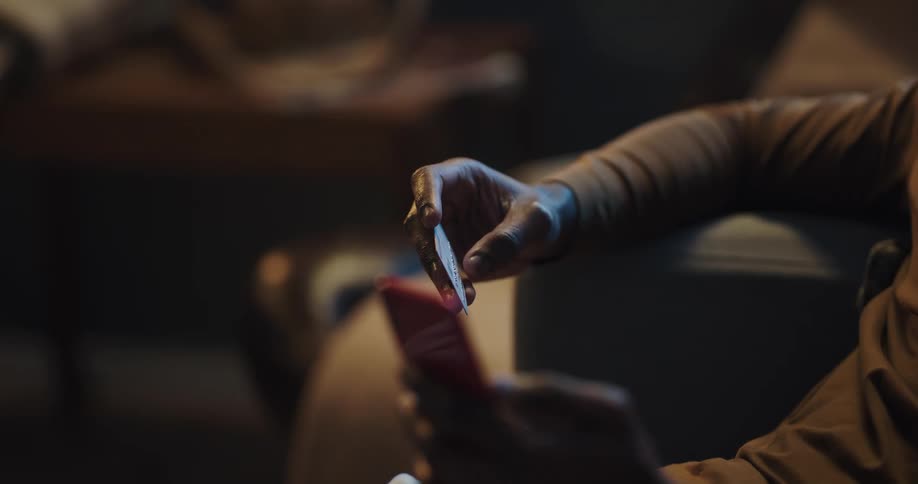 A man sits at home and scrolls on his cell phone and holds a credit card in the other hand