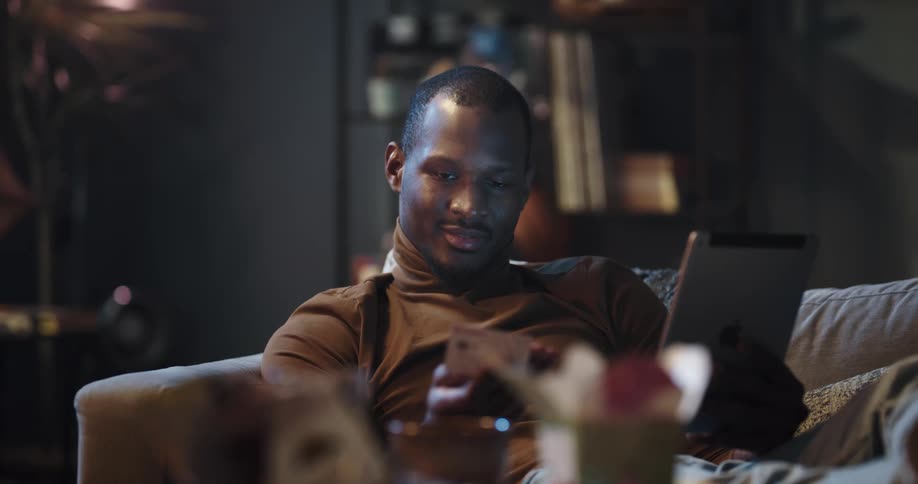 Black man sitting on the couch typing his credit card information on the tablet