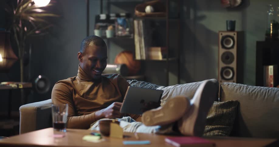 Man sits on a sofa and laughs and smiles as he looks at a tablet
