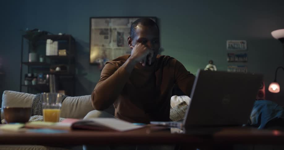 A man sits on a sofa in the living room while working on a laptop surrounded by items
