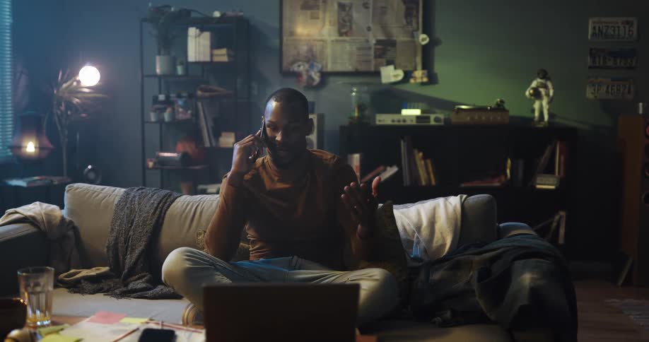 Man sits cross legged on couch talking on phone while viewing laptop
