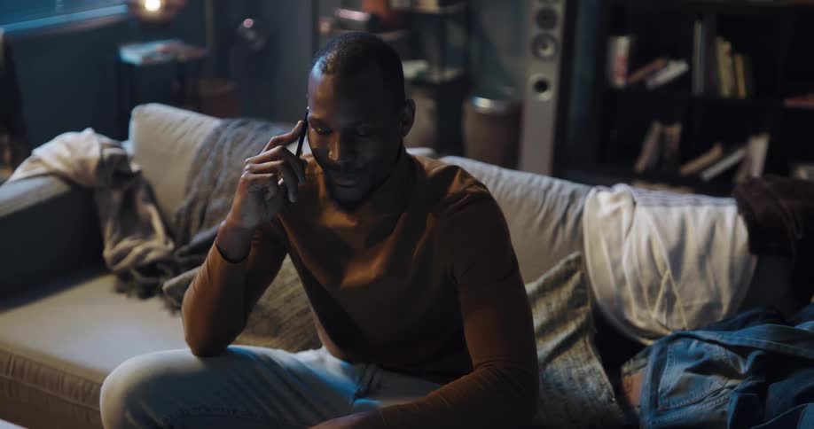 A man laughs while sitting on the couch and talking on the phone