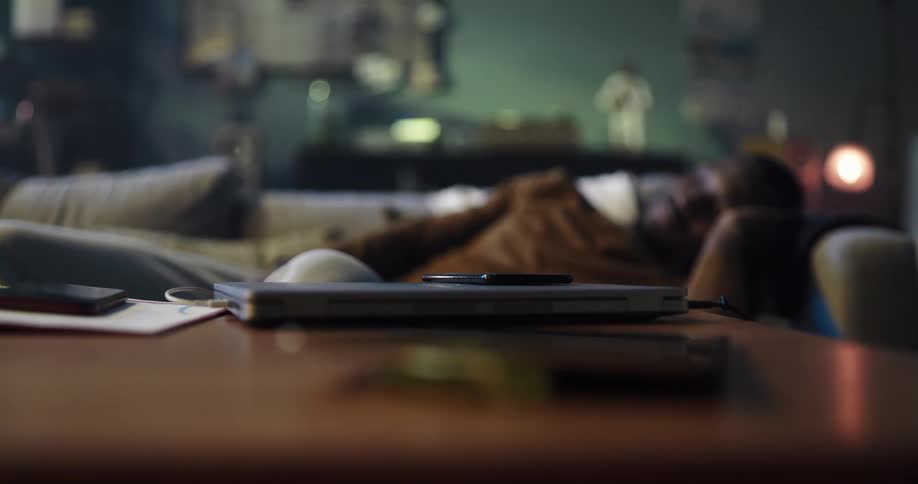 A closed laptop with smart phone on top sits on a wooden table near a man sleeping on a couch