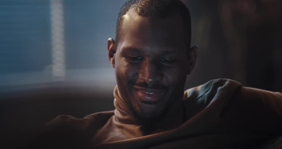 Man with beard and brown turtleneck sweater sits on couch using smartphone and smiling in living room at home
