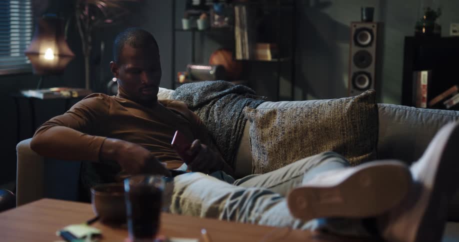 Young man laying on the couch in the living room takes a credit card out of his jean pocket to make a purchase on his phone