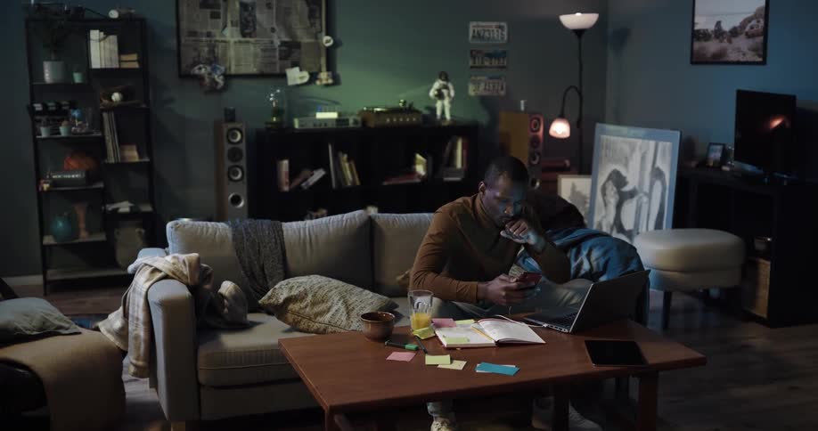 Man with brown turtleneck sweater and jeans sits on couch using smartphone and laptop in the living room