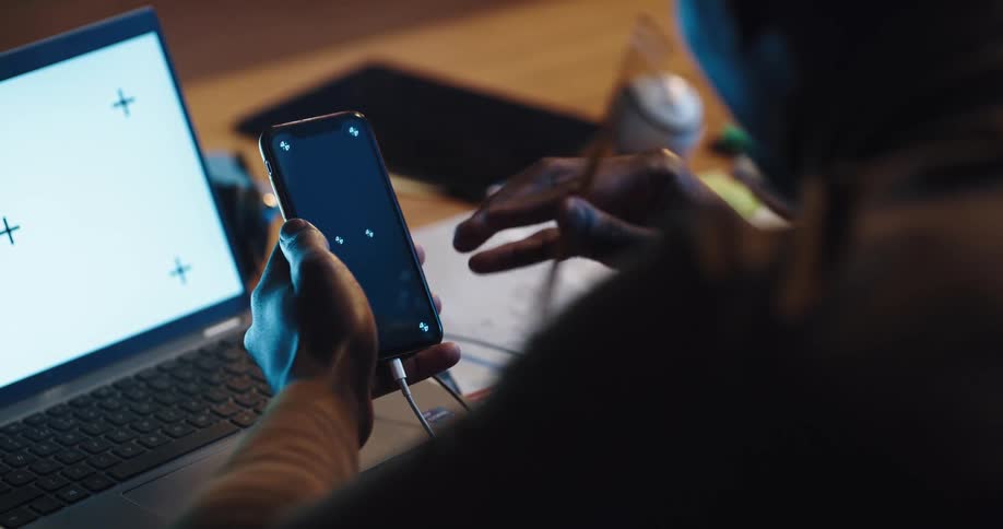 A man taps the screen of smartphone connected to a laptop via usb while sitting at a desk