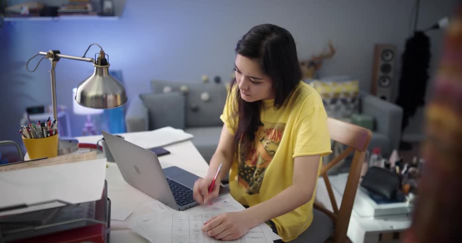 Woman with dark hair and yellow t shirt sits at table using laptop and writing down in notebook in room
