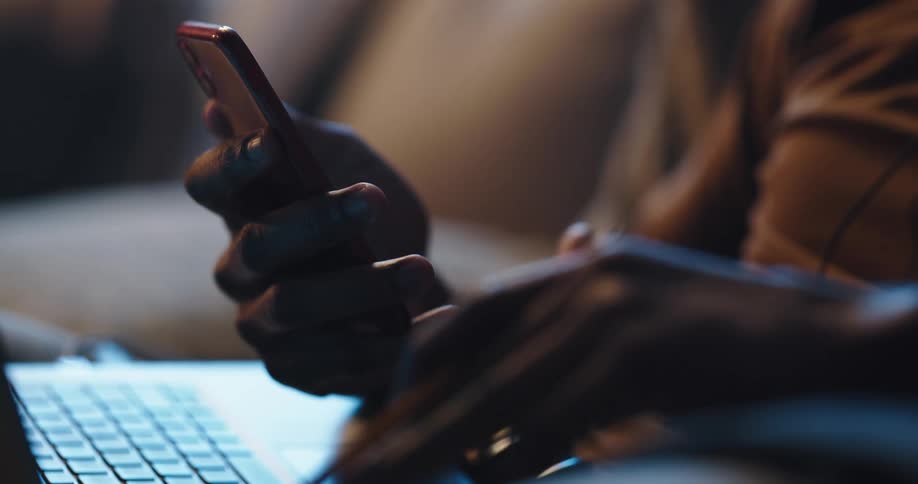 Man with brown sweater sits on couch using smartphone and laptop in living room at home