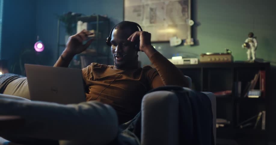 Man relaxing on the couch in the living room puts on headphones while talking on a video call through a laptop