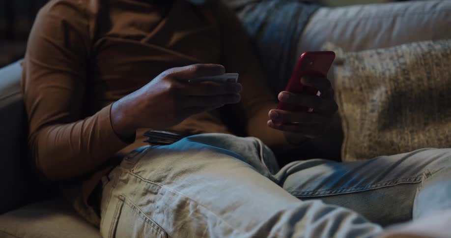 Man reclines on a sofa while holding a cellphone and reaches into his pocket to get a credit card