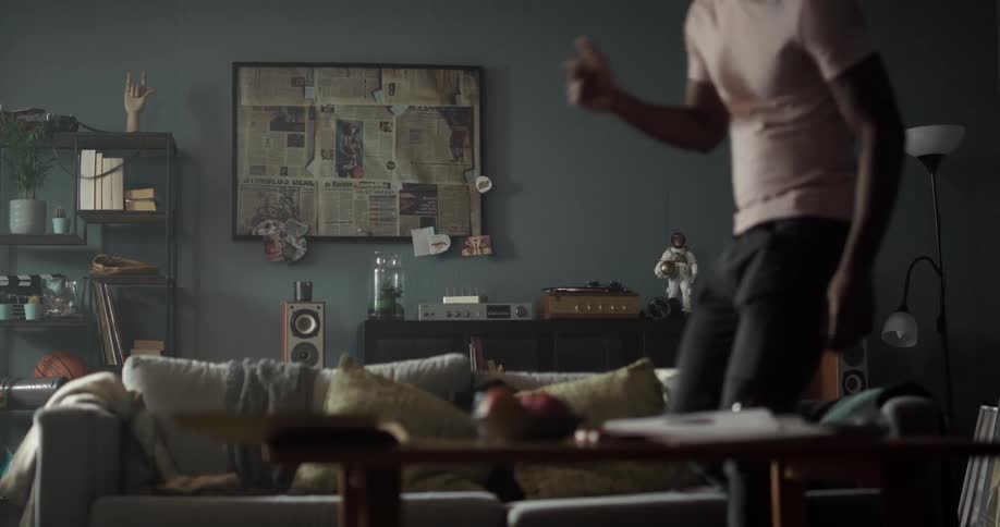 Black man putting the needle on a vinyl record on a record player and starting to dance to the sound of the music while walking towards the couch and then sitting down