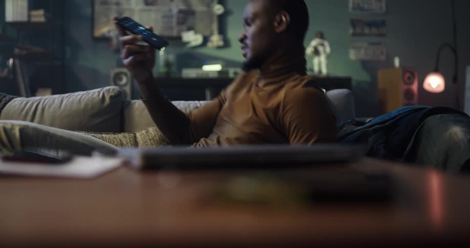Man with brown turtleneck sweater lies on couch and answers call on smartphone in living room at home