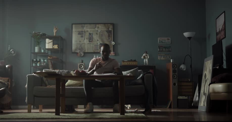 Man turns away from a shelf with electronics on it and dances to the sofa then sits down and puts one foot up on a knee