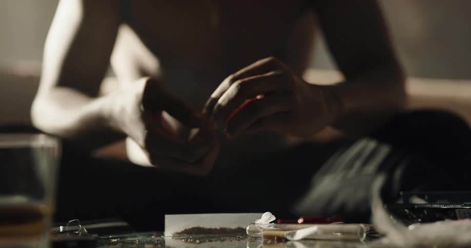 Black man sitting on the couch in front of a table with drugs and a drink rolling a joint