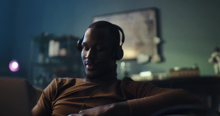 Man wearing a headset with a microphone sits on a sofa and talks while looking at a laptop