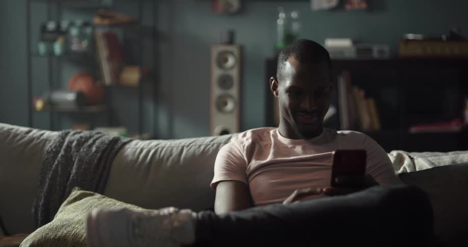 A man is sitting indoors and texting on a cellphone