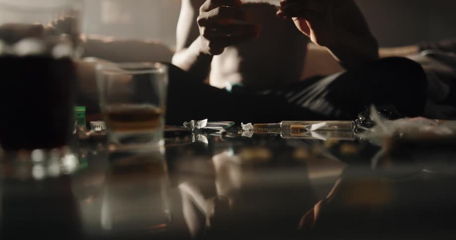 Shirtless man sitting on the couch in front of a glass top table filled with drinks and drugs rolling a joint