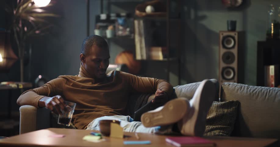 A man resting on a sofa drinks from a cup while watching a video on his computer tablet