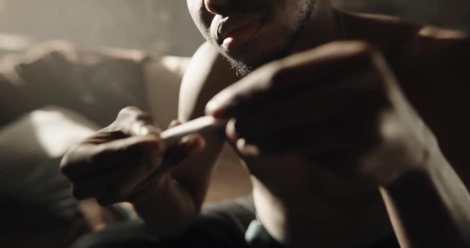Shirtless black man sitting on the couch rolling a joint and licking the rolling paper to seal it