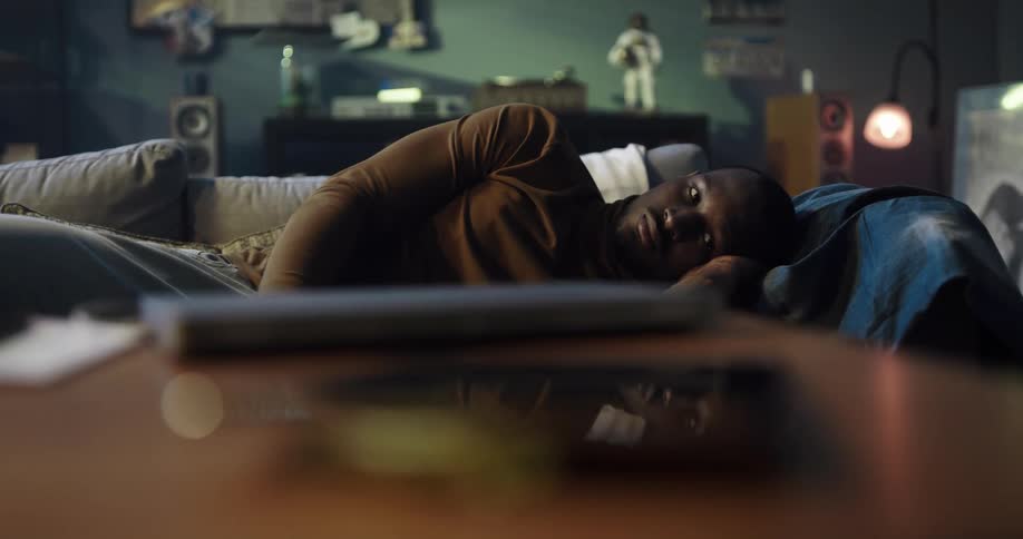 Black man lying on the couch with a bored and thoughtful facial expression sitting up to talk to the phone