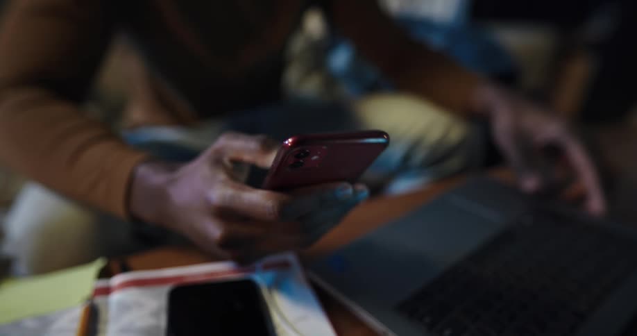 Man with brown turtleneck sweater and jeans sits on couch using smartphone and laptop in living room at home