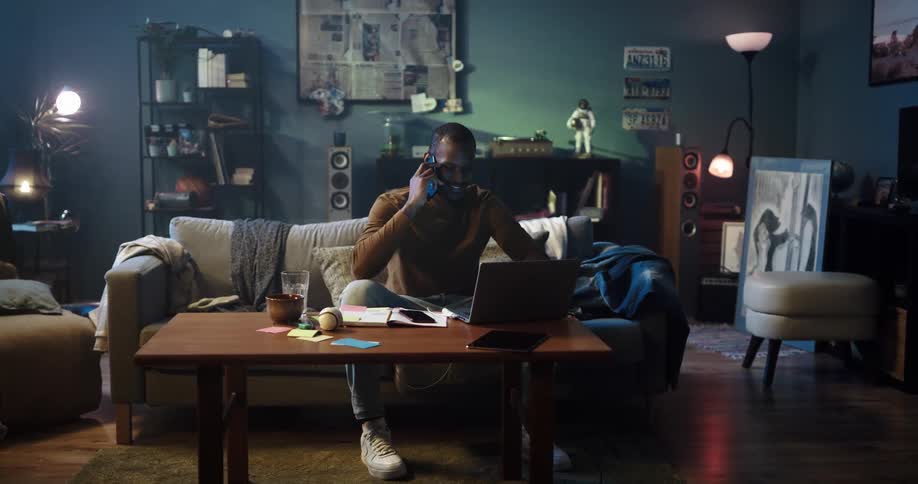Man sitting on the couch at home uses a laptop while talking on the phone
