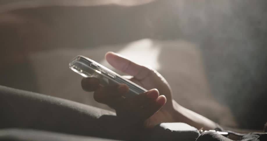 A man sitting in bed lights a cigarette and scrolls on his cell phone as he smokes