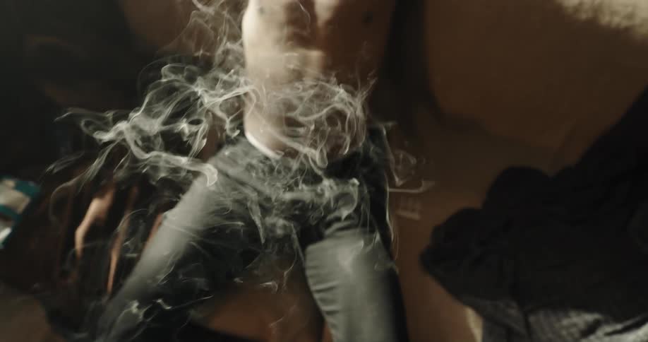 Young man laying in a couch at home smokes a cigarette watching the smoke rise at morning