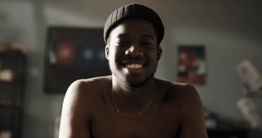 Shirtless man with black wooly hat and necklace faces the camera with eye contact smiling