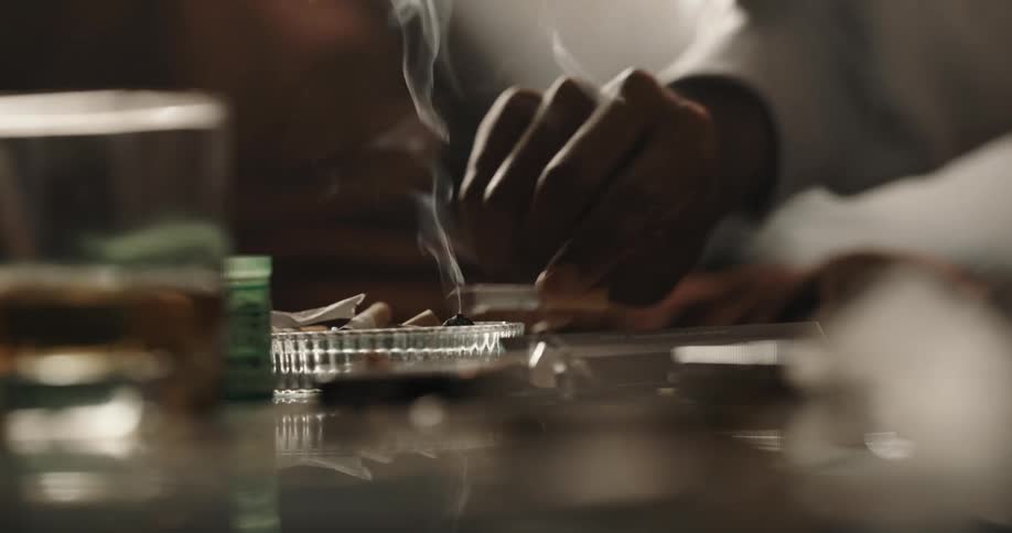 A smoldering cigarette is picked up from the edge of an ashtray before being put down again