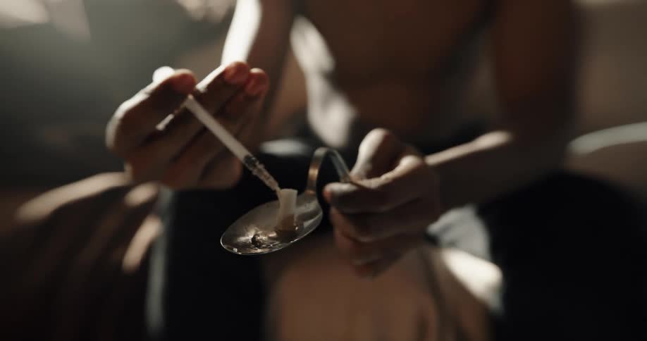 A seated male dips a syringe into a liquid that he is holding in a spoon