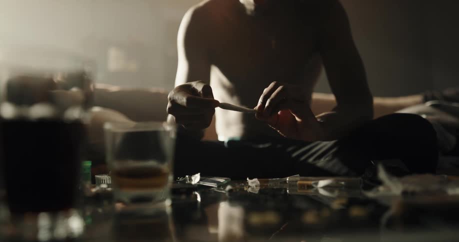 Shirtless black man sitting on the couch rolling a joint with many beverages and drugs in the table in front of him