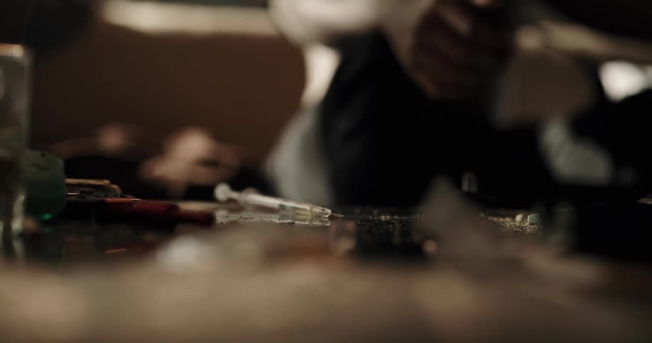 Shirtless black man sitting on the couch tying a belt around his arm to inject a syringe with drugs